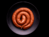 Longaniza selecta cruda en forma de espiral, colocada sobre un plato negro con fondo oscuro tipo estudio fotográfico.