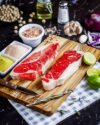 Dos cortes de carne tipo New York nacional sobre tabla de madera, acompañados de condimentos, limón, ajo y cebolla morada.