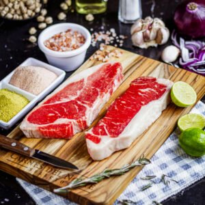 Dos cortes de carne tipo New York nacional sobre tabla de madera, acompañados de condimentos, limón, ajo y cebolla morada.