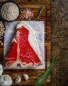 Corte de carne T-Bone nacional crudo sobre tabla de madera, acompañado de sal rosa, romero, ajo y especias.